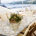 Luxurious yacht deck with a marble dining table, wooden chairs, floral centerpiece, and a scenic ocean view.