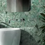 Modern bathroom featuring a glossy white vessel sink, textured green stone wall, silver faucet, mirror, and tall plant.
