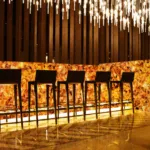 Luxurious bar scene with dark chairs lined against an illuminated amber-colored bar under elegant ceiling lights.