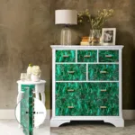 White dresser with green marble-patterned drawers, a lamp, books, and vase beside a white side table with flowers.