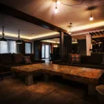 Cozy modern living room with dim lighting, dark sofas, a patterned wooden coffee table, and industrial ceiling lights.